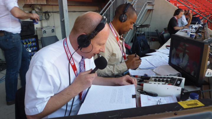 John Roder holds a microphone to his face while wearing over-ear headphones and looking at handwritten notes, in a seating area of a football ground