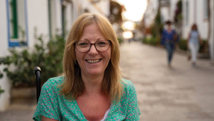 A white woman in her 50s wears glasses, a turquoise floral dress and uses a wheelchair 