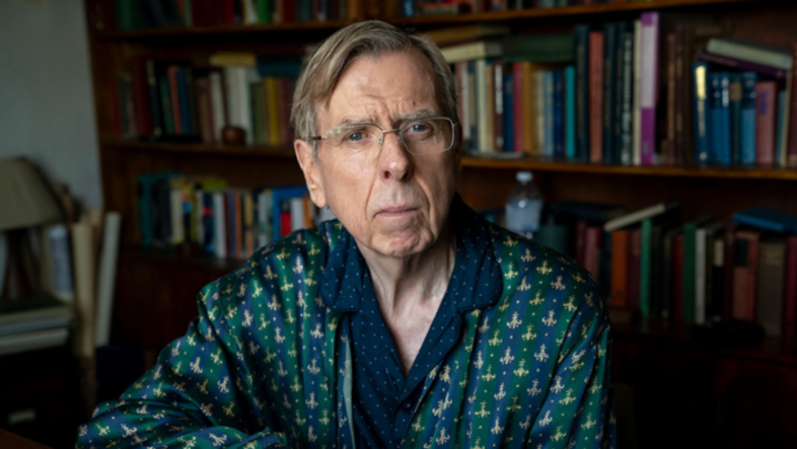 Timothy Spall, a man in his mid sixties, sits in front of a large fully packed bookcase, in his blue polkadotted pyjamas and a green and navy striped silk dressing gown. He wears rectangular glasses.