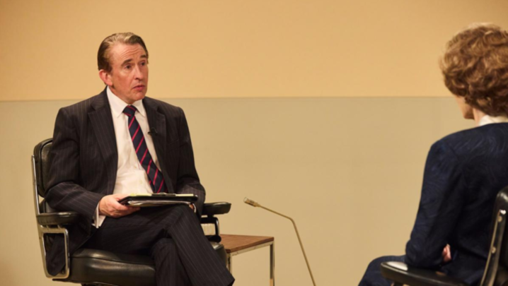 In a TV studio, Steve Coogan looks at Margaret Thatcher, all of whom we can see is the back of her head