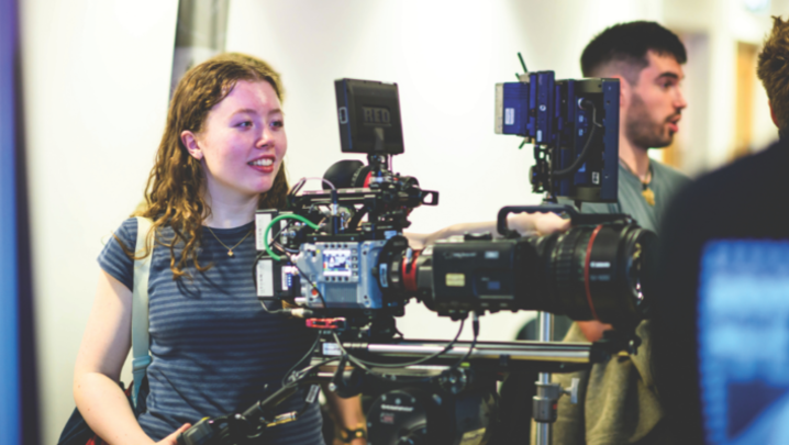 A young woman in a stripy top operates a large, industry-standard video camera