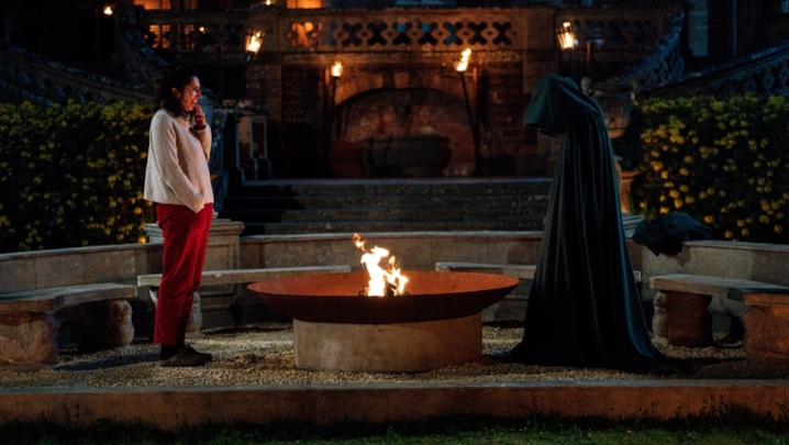 A white woman with dark hair stands by a firepit outside a castle and opposite a fully cloaked person