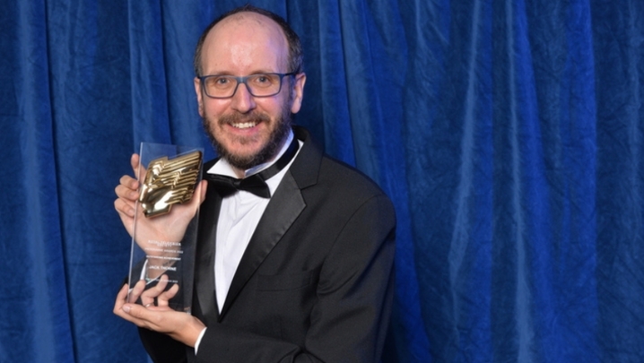 A white man with brown hair, moustache and beard wearing black tie, sits down and holds up a trophy against a backdrop of blue curtains