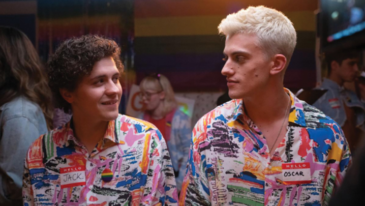 Dylan Llewellyn and Jon Pointing stand in a party, looking at each other in matching multi-coloured shirts, wearing name tag stickers 