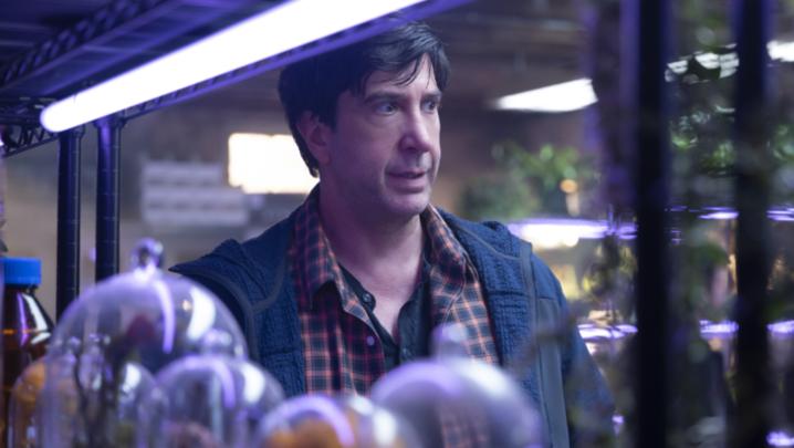 David Schwimmer, a white man in his fifties, looks stressed in a dark laboratory, surrounded by plants and bell jars