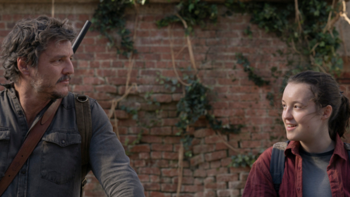 Pedro Pascal, a Chilean-American man in his forties, and Bella Ramsey, a person in their late-teens in the image, stand side by side, looking at each other in front of a brick wall