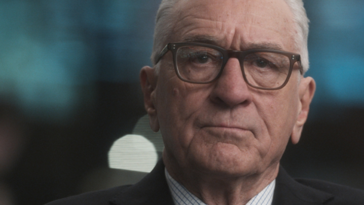 Robert De Niro, a white man in his eighties, is unsmiling in glasses and a suit, a blue and dark backdrop blurred behind him