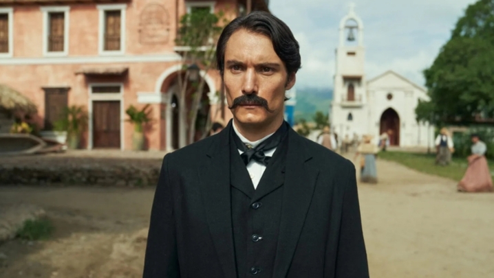 Aureliano looks stern dressed in a formal suit in the fictional town of Macondo, with a Church pictured behind him