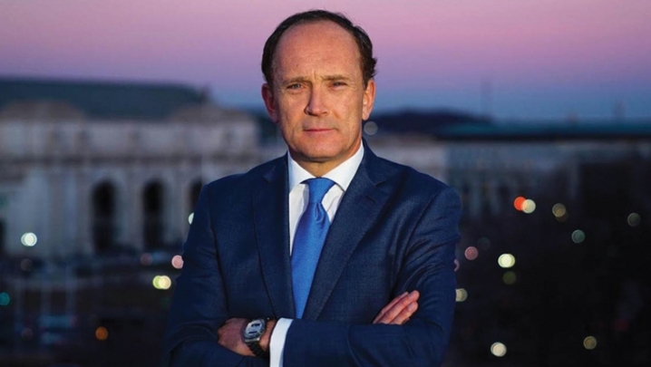 A white man with dark hair wears a navy suit and stands with his arms crossed against a purple sunset backdrop