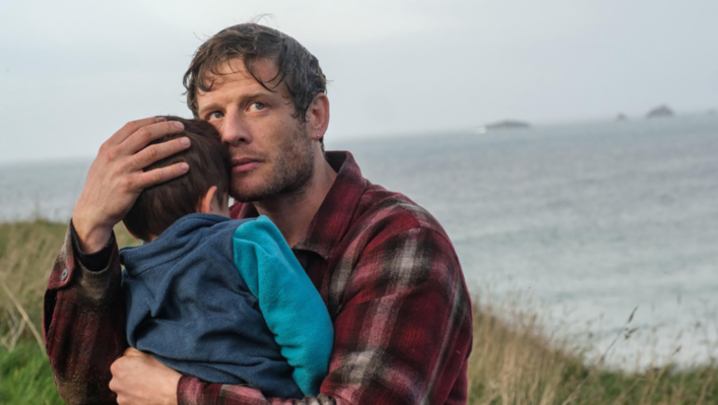 James Norton cradles a child, standing close to the sea