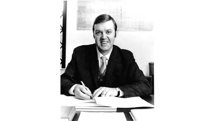 A black and white photo of a white man in a suit sitting at a desk and writing on a notepad