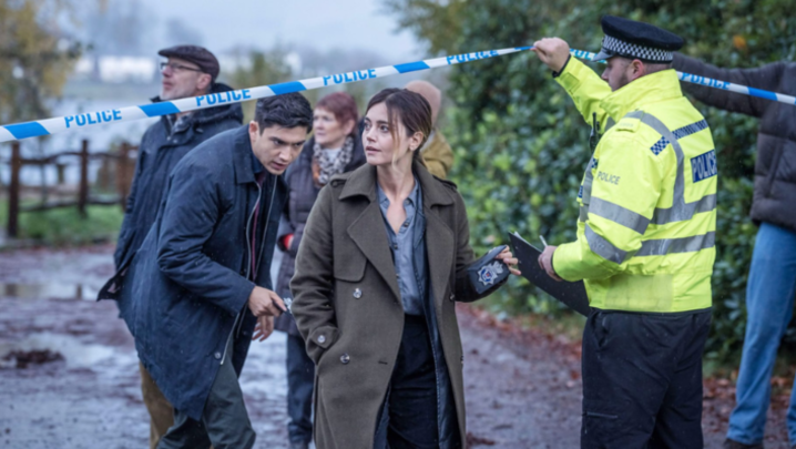 Archie Renaux as Hitch and Jenna Coleman as Ember walk under police tape being held up for them by an officer in a hi-vis tabard
