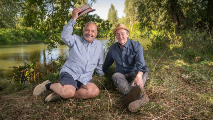 Bob Mortimer and Paul Whitehouse sit next to each other by a lake, looking into the camera and smiling, with Mortimer taking his hat off in friendly greeting