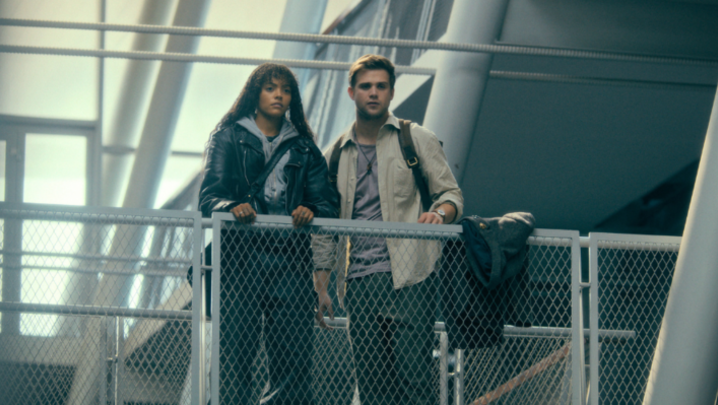 Quintessa Swindell, a biracial woman in her 20s, and Leo Woodall, a white man in his late 20s, stand together on a walkway indoors