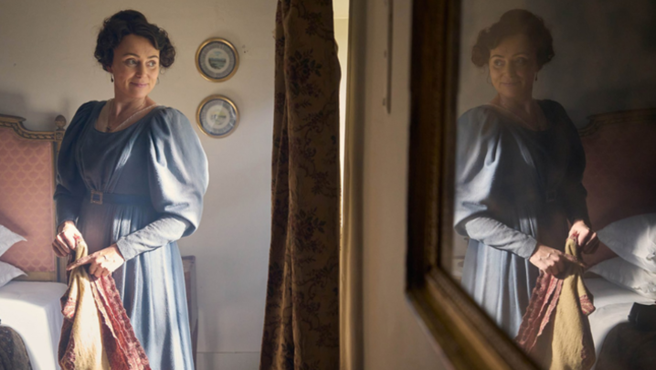 Keeley Hawes, a white woman in her forties, holds a sheet while looking out the window, smiling and wearing a blue 19th century everyday-wear dress