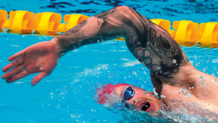 A swimmer doing some kind of front crawl looks to the side, half his face submerged in the water, his hand poised to re-enter the water