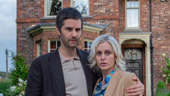 Denise Gough, a white woman in her forties, and Jim Sturgess, a white man in his forties, stand arm in arm outside a large-ish suburban red brick house, unsmiling