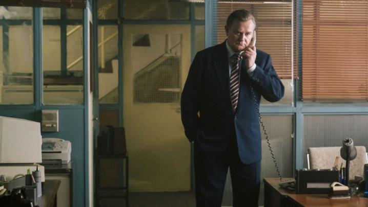 Hugh Bonneville, a white man in his sixties, stands in an office on a phone with a cord, looking concerned