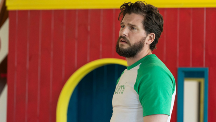 Kit Harington stands indoors, looking crestfallen in a white and green t-shirt with the logo for Lumi, an in-world green energy company 