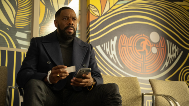 Colman Domingo, a black man in is fifties, sits indoors in a navy blue peacoat-style waist-length jacket, holding a phone and small piece of paper