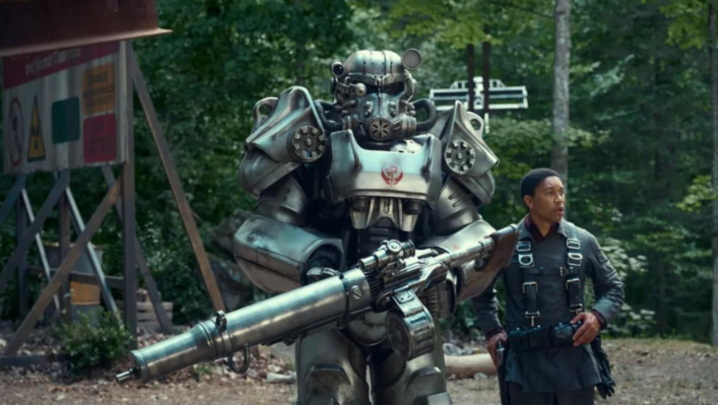 Two people walk in a forest, one in a large chrome mec suit