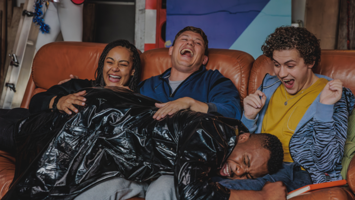 A group of four friends sit on a rust coloured sofa, laughing as one of them lies on their laps