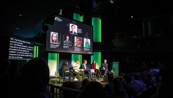 A panel of 3 men and two women sit on a stage in front of a screen with their pictures and names