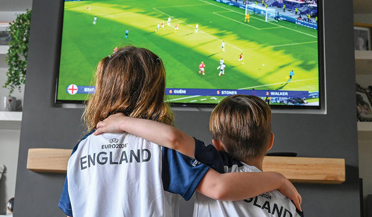 Two kids wearing England football shirts watch England play in Euro 2024 on TV 