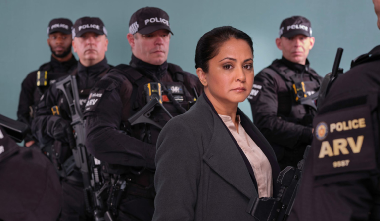 A woman in a grey coat stands in front of a group of armed police