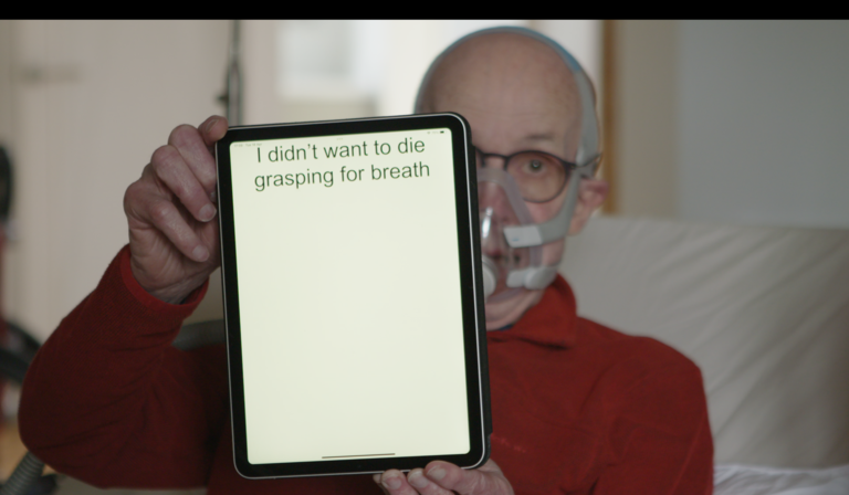 A man with breathing apparatus holds an iPad to camera, with text on the screen reading 'I didn't want to die grasping for breath'