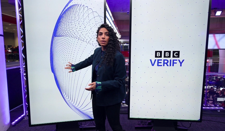 A woman presents in front of LED screens, one of which has text reading 'BBC Verify'