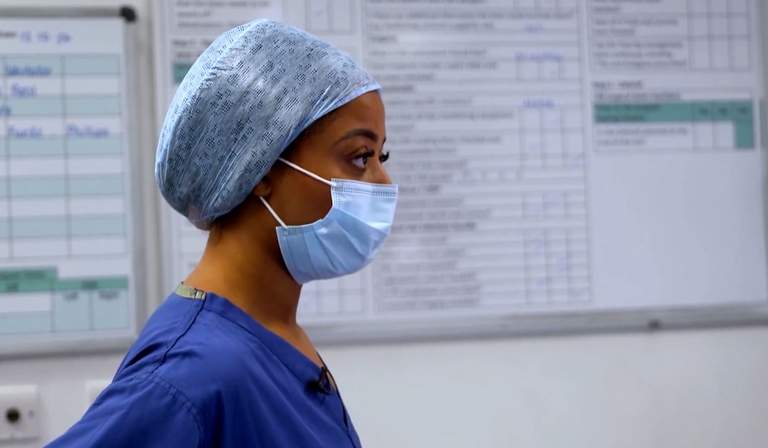 Cree stands in a hospital corridor, wearing a face mask, blue scrubs and a blue hat containing her hair