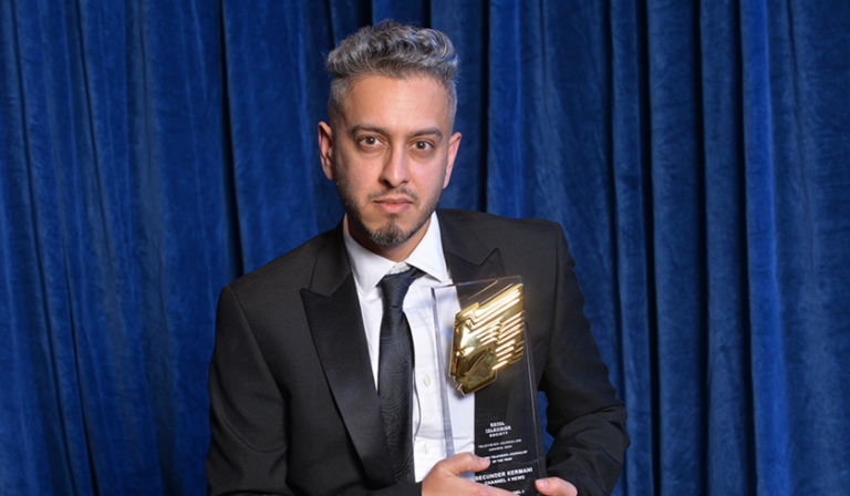 Secunder Kermani sits in a suit holding an RTS Award, looking into the camera in front of a blue curtain backdrop