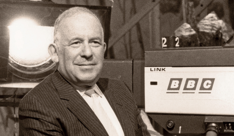 In a black and white image, Paul Fox sits next to a camera with the 1980s version of the BBC logo