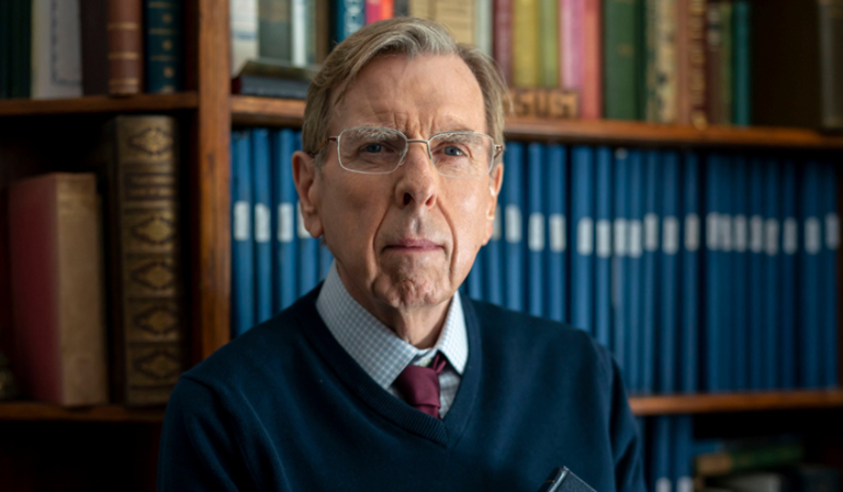 Timothy Spall stands in front of bookshelves, wearing glasses and looking pensive