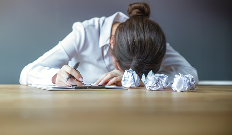 A woman sits with her head on a desk, next to scrunched up bits of paper