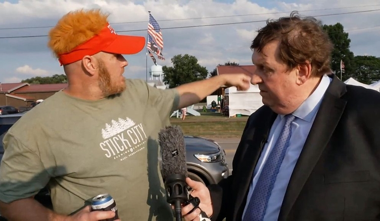 A journalist in a suit interviews a man in a t-shirt and hat, who points off camera