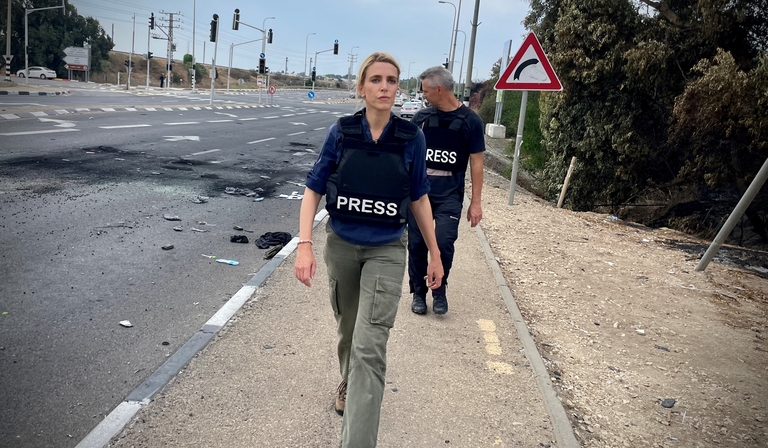 A woman walks down a street wearing a body armour vest labelled 'Press'
