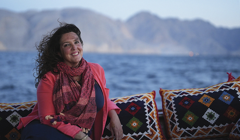 Bettany Hughes is pictured on an outdoor sofa overlooking a mountainous bay