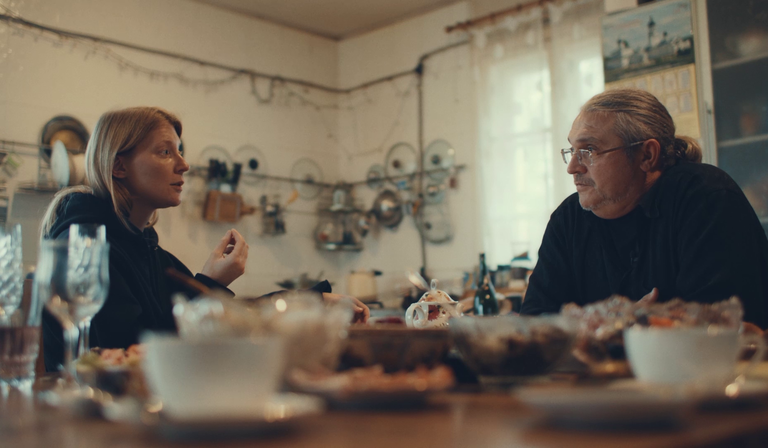 A man and a woman sit across from each other around a kitchen table, discussing something