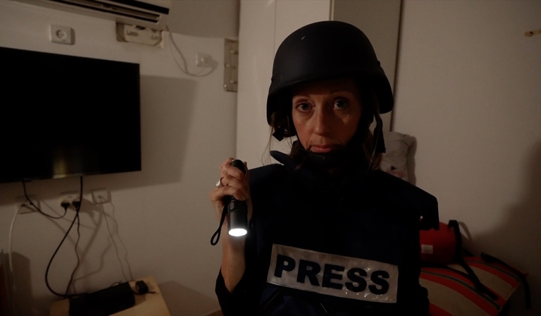 A news reporter stands in a house, holding a torch and wearing a body armour helmet and vest, the latter of which reads 'Press' 