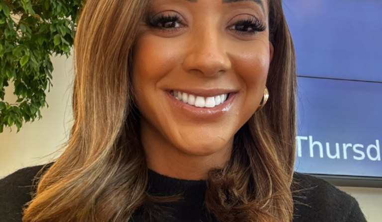 A woman with shoulder-length, light brown hair is smiling. She is wearing a black top and gold hoop earrings. The background includes a partial view of a large screen and a green potted plant.