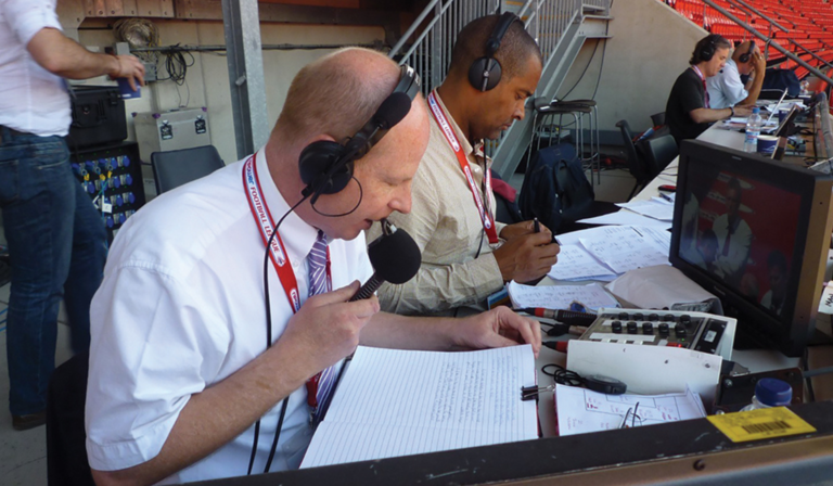 John Roder holds a microphone to his face while wearing over-ear headphones and looking at handwritten notes, in a seating area of a football ground