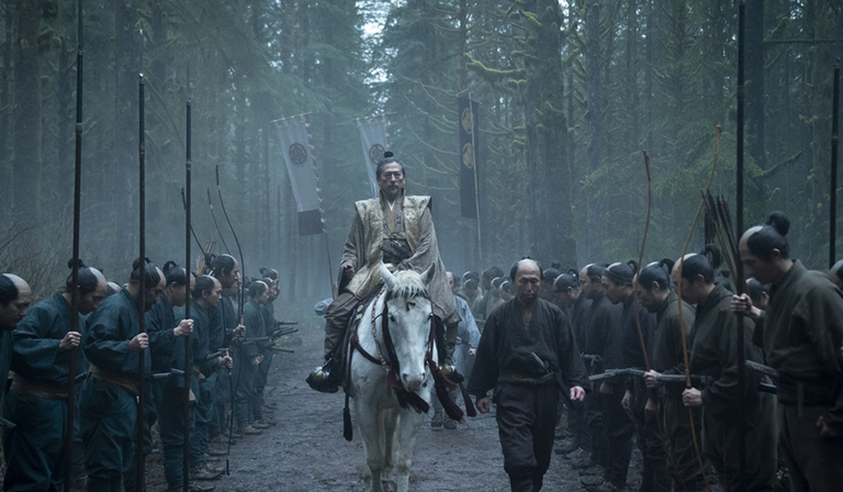 A military general on horseback trots past his troops in a forest as they bow their heads in deference