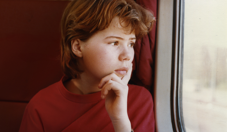 A young girl looks out the window, sat on a train. The picture is grainy, suggesting it was taken on film