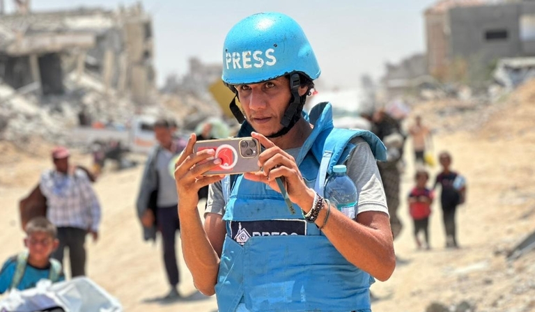 Mohammed films something with a phone, wearing a blue bulletproof vest and helmet. The helment has the word "PRESS" written on it in large white lettering