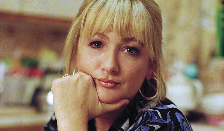 A headshot of Caroline Aherne, a white woman with blonde hair wearing a blue and black patterned shirt