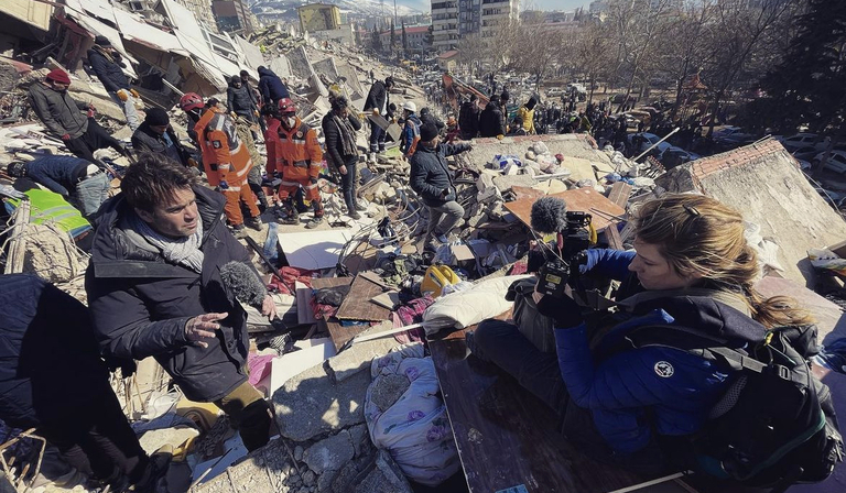A news reporter delivers a piece to camera, operated by a camerawoman, the two of them both stood on rubble