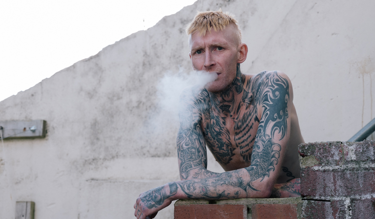 A shirtless man whose chest, arms and hands are covered in tattoos blows smoke out of his mouth while looking into the camera