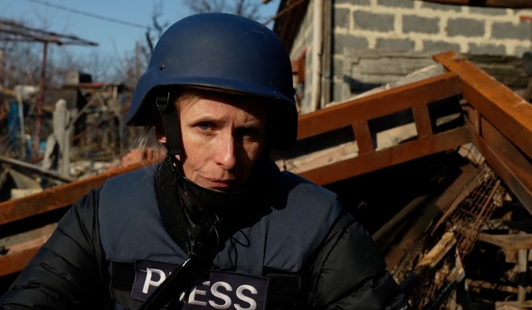 Orla Guerin stands in front of rubble in a body armour helmet and vest, the latter of which has 'Press' written on it